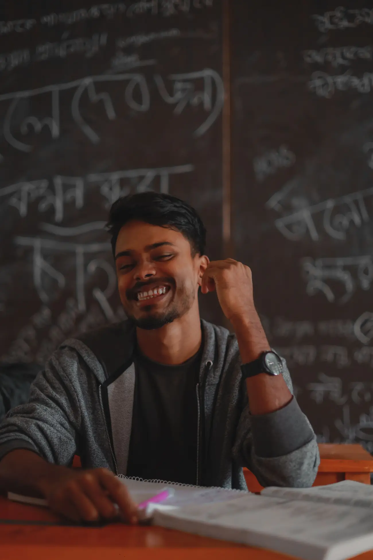 A young man smiling at his desk in front of a chalkboard